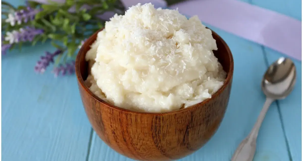 Una ciotola di legno piena di cremosa crema al cocco, guarnita con cocco grattugiato. Accanto, un cucchiaino d'argento e fiori di lavanda.