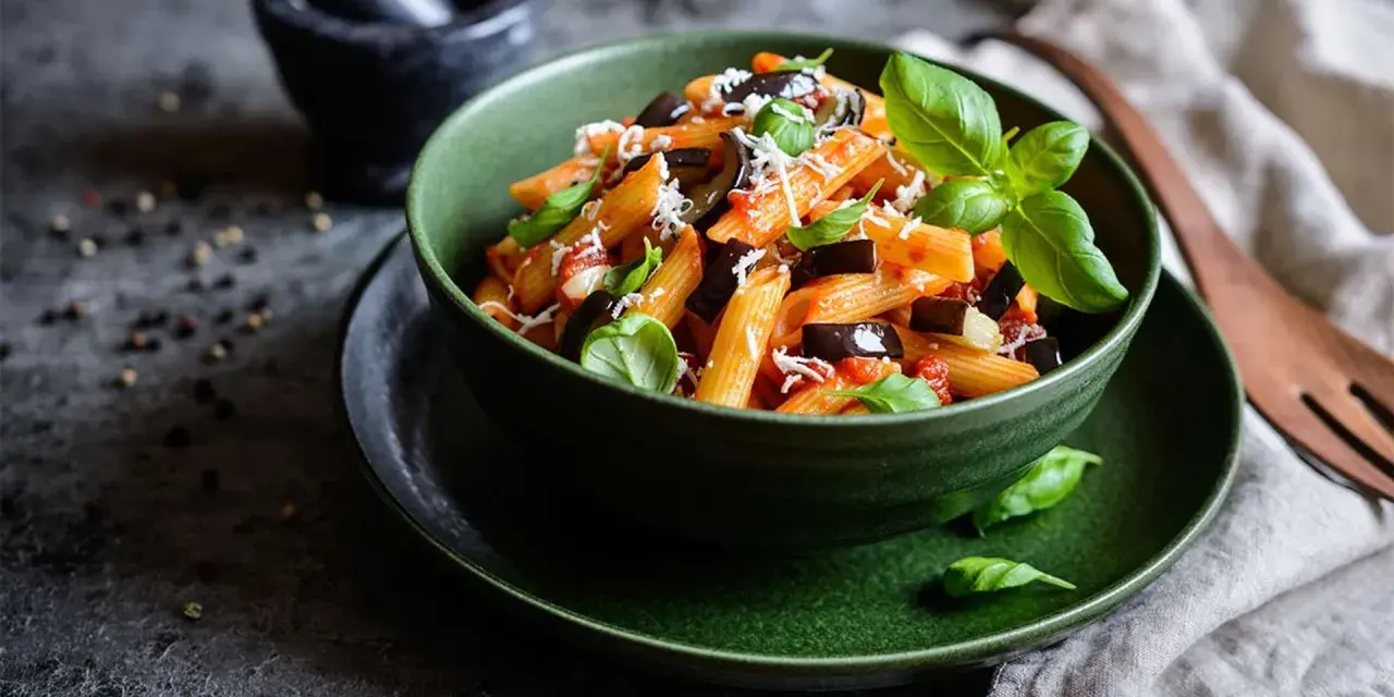 Un plato de pasta alla norma, con penne, berenjena, salsa de tomate y albahaca fresca, espolvoreado con queso rallado.
