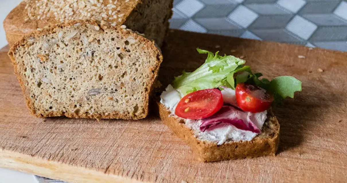 Keto chleb majonezowy przepis: kromka z serkiem, pomidorkami i sałatą obok bochenka z ziarnami.