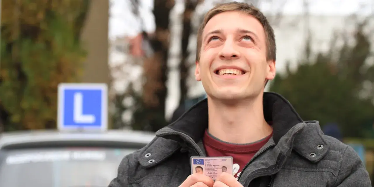 Szczęśliwy młody mężczyzna trzyma prawo jazdy kategorii B, z niebieskim znakiem "L" na dachu samochodu w tle.