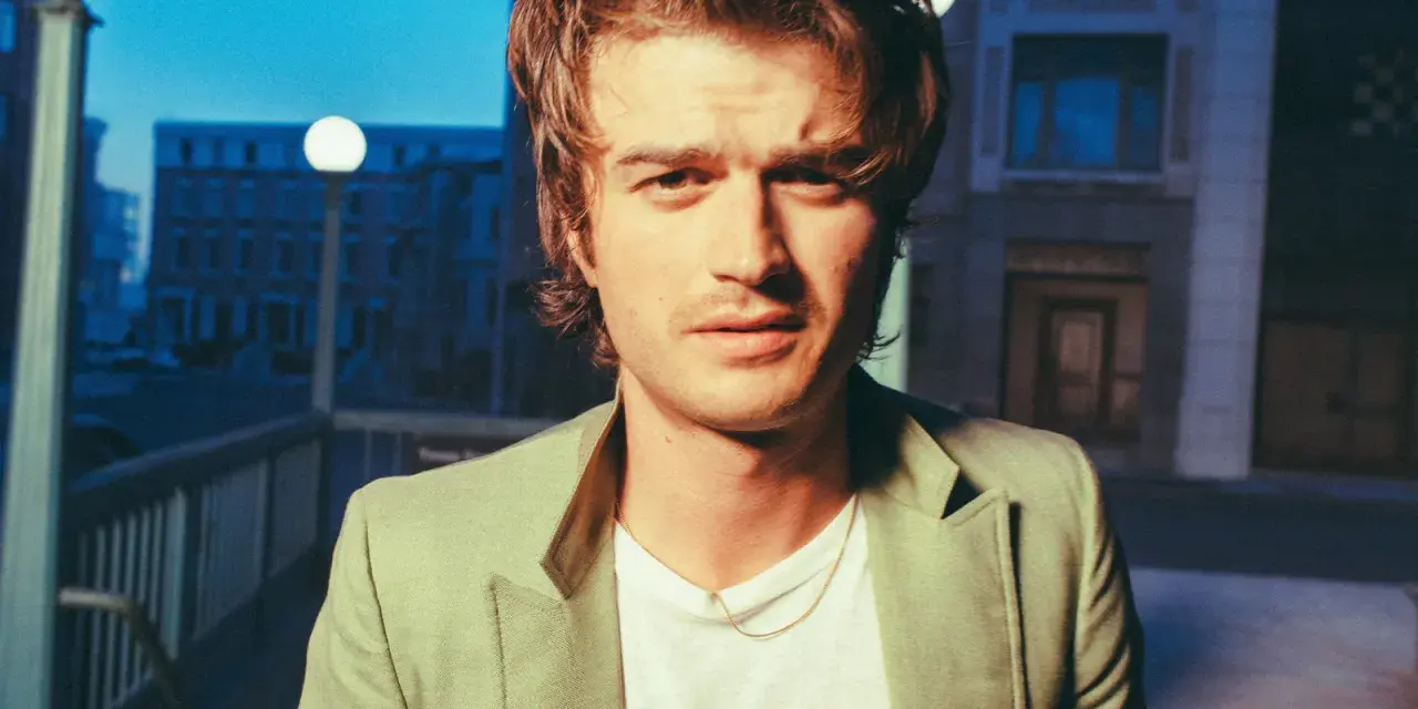 Joe Keery w jasnej marynarce, z naszyjnikiem, na tle nocnego miasta.