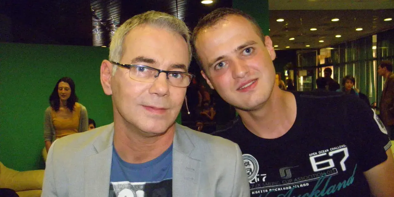 Tomasz Kammel i młody fan pozują do zdjęcia. Tomasz Kammel w szarym garniturze i niebieskiej koszulce, a obok niego młody mężczyzna w czarnej koszulce z napisem "Auckland".
