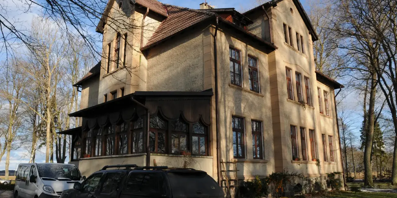 Dawny pałac Poraj, z charakterystyczną werandą i drzewami w tle, otoczony zimowym krajobrazem.