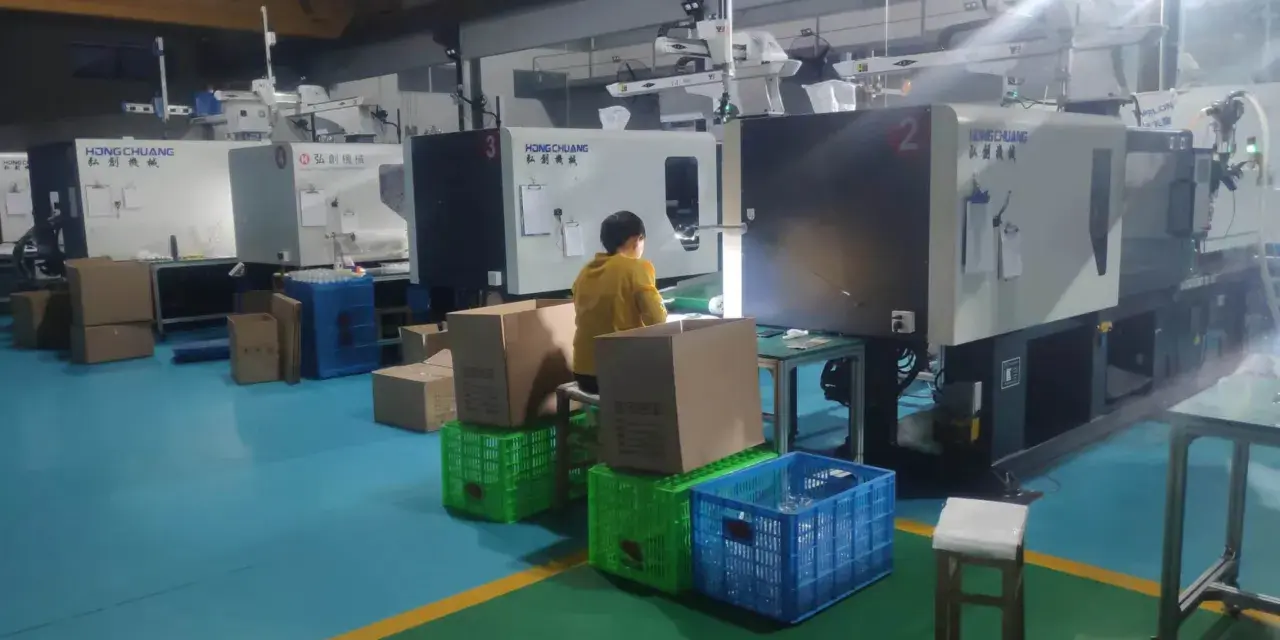 A person in a yellow jacket works at a station with boxes and crates, surrounded by injection molding machines, indicating low volume production.