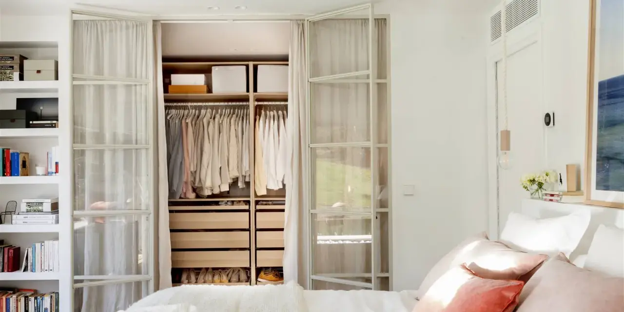 Dormitorio con vestidor integrado. Ropa organizada en armarios abiertos y estanterías. Cama con cojines rosas y manta blanca.
