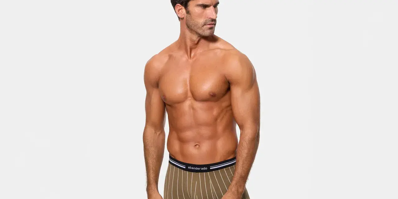 Hombre musculoso posando con calzoncillos de poliamida abriga, con rayas verticales blancas sobre fondo verde oliva.