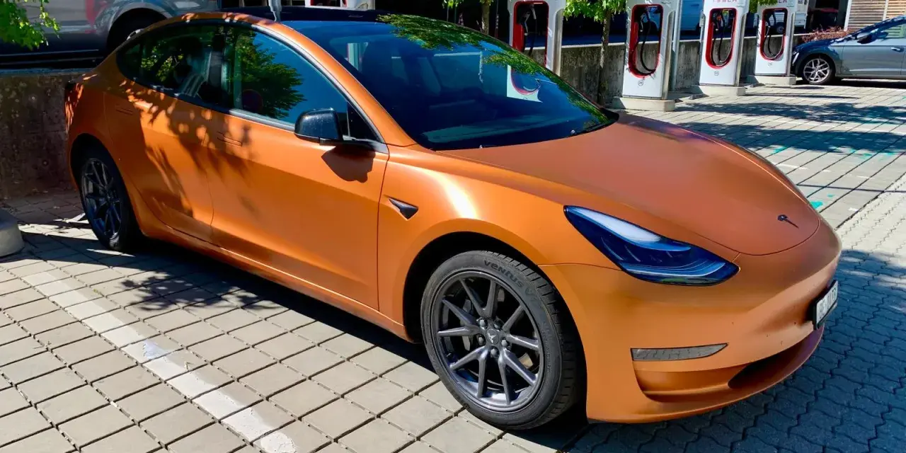 Pomarańczowa Tesla na tle ładowarek Supercharger.