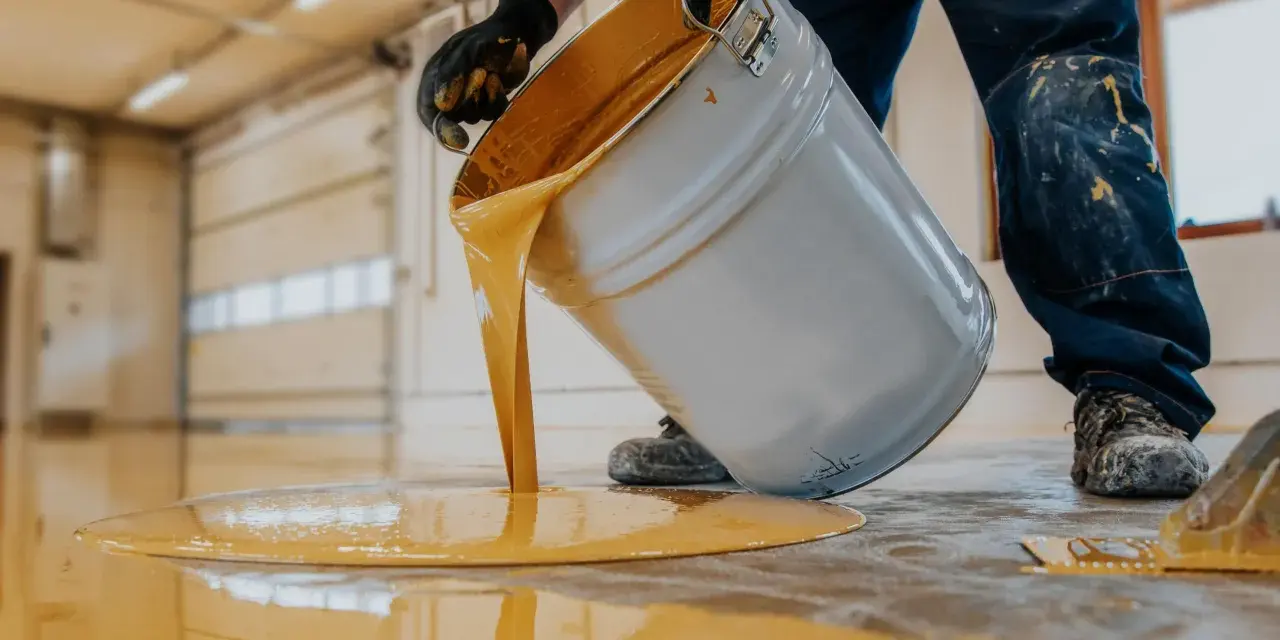 Hombre con guantes vierte pintura para suelo de cemento de color amarillo brillante de un cubo.