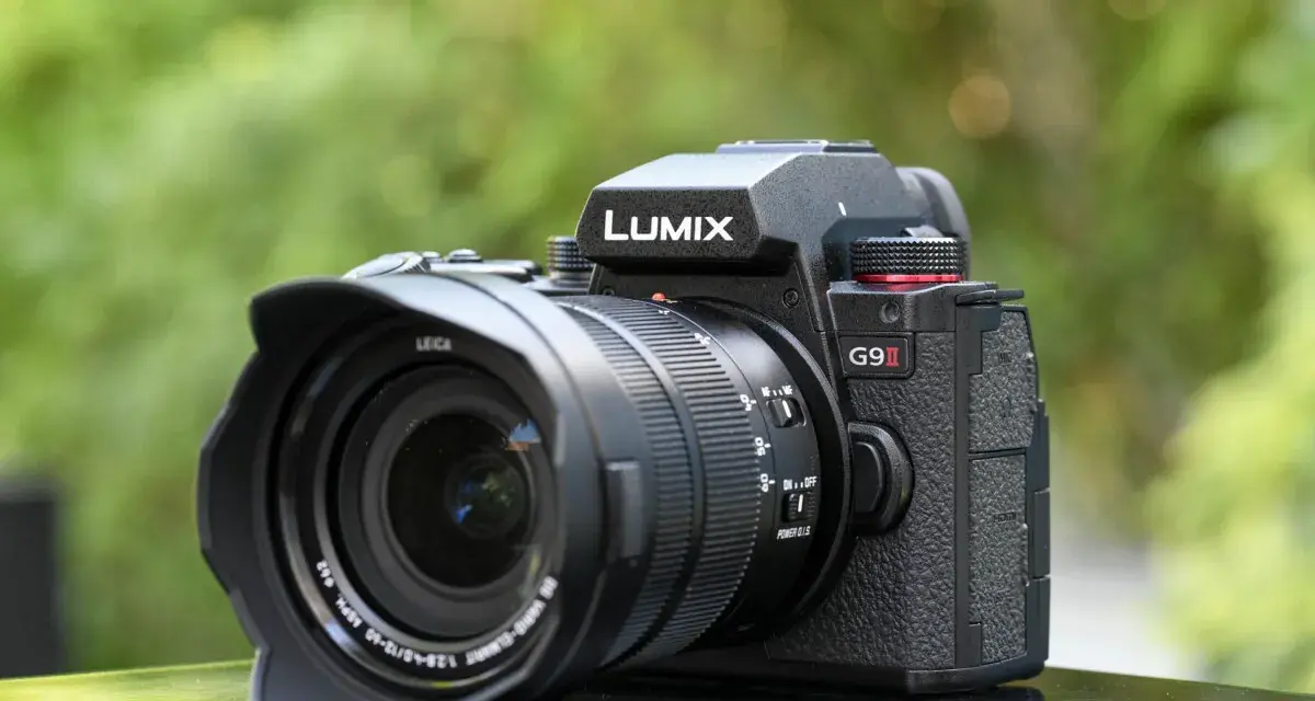 Un appareil photo noir Lumix G9 II avec objectif Leica est posé sur une surface réfléchissante, avec un arrière-plan flou de verdure.