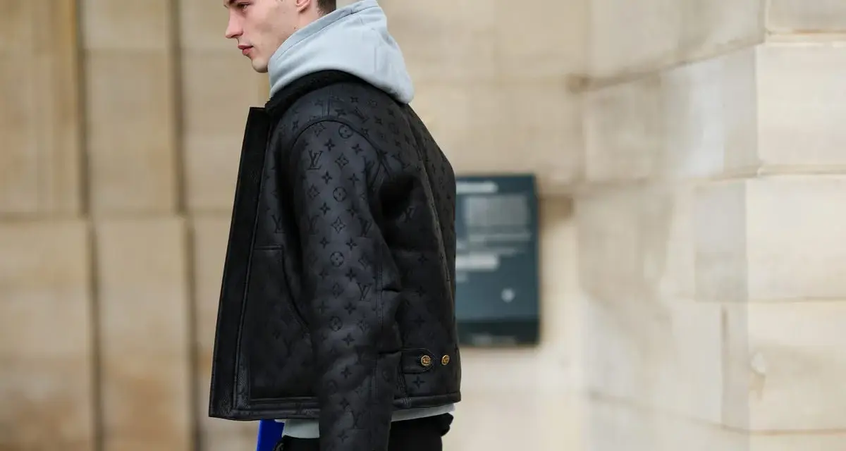 Uomo con un elegante outfit con giubbotto di pelle nero con monogrammi, felpa grigia e pantaloni scuri.