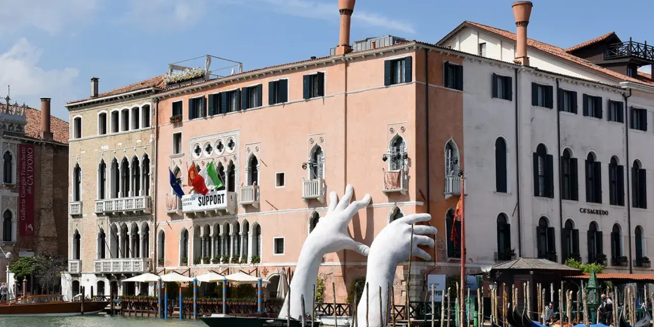 Enorme installazione di mani bianche emerge dal Canal Grande, davanti al museo arte contemporanea Venezia. Gondole ormeggiate.