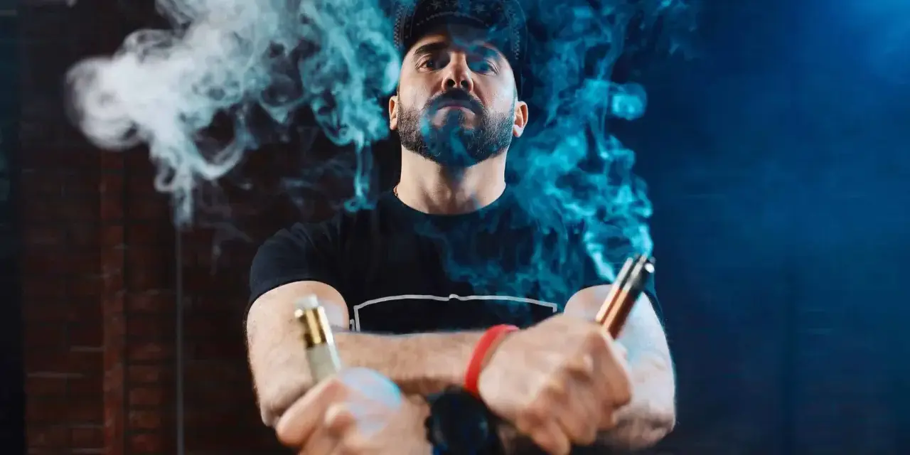 Hombre con barba y gorra, rodeado de vapor azul, sostiene dos vapers, uno dorado y otro cobre. La diferencia entre 0.8 y 1.2 ohm se siente en la calada.