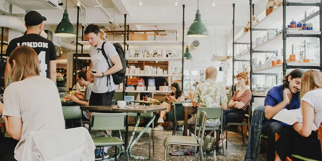 Gente en un café con mesas verdes y estanterías llenas de objetos. El **ruido restaurante** es palpable con las conversaciones y el movimiento.