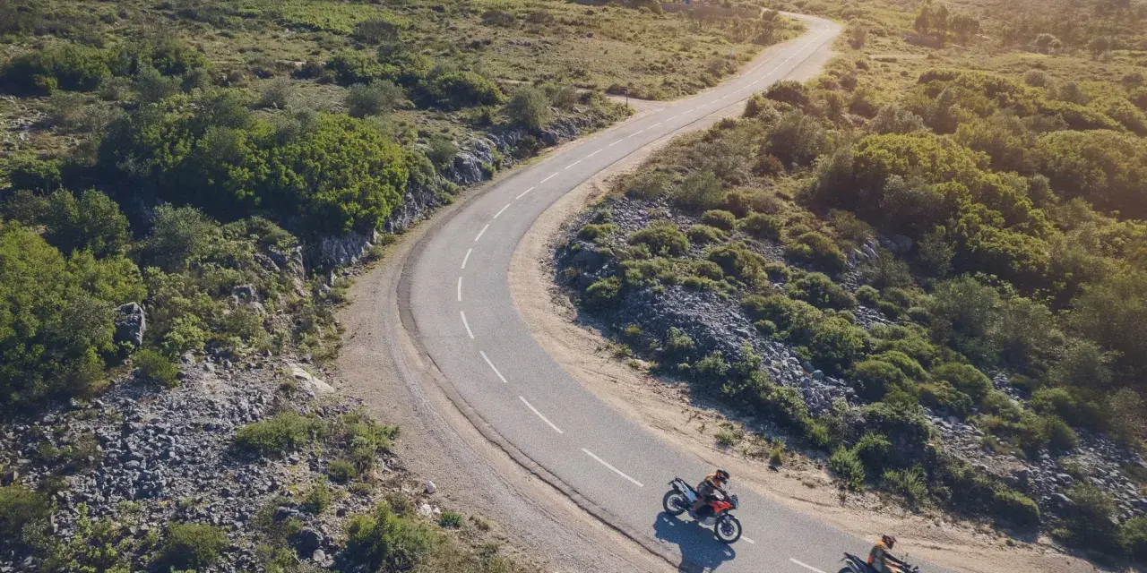 Due moto KTM sfrecciano su una strada tortuosa tra colline verdi. Un'avventura che celebra il KTM Connect.