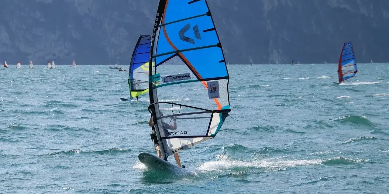 Osoba na desce windsurfingowej z niebieskim żaglem na wzburzonym morzu.