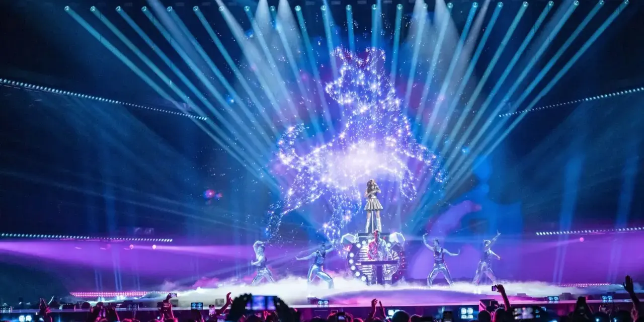 Wokalistka na scenie Eurowizji, otoczona tancerzami i efektami świetlnymi. Widownia filmuje występ, zastanawiając się, ile krajów w finale Eurowizji.