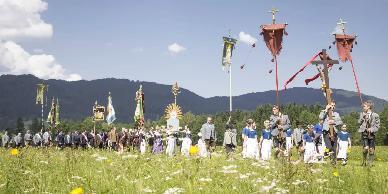 Menschen in Tracht und Uniformen nehmen an einer Prozession teil, die den Fronleichnam feiert. Fahnen und ein Kreuz werden getragen.