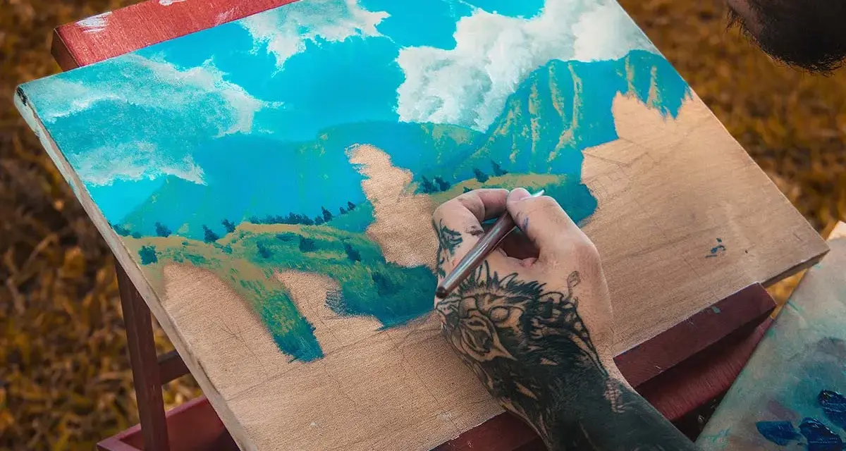 Un artiste tatoué est en train de peindre à la gouache sur toile un paysage montagneux sous un ciel bleu et nuageux.