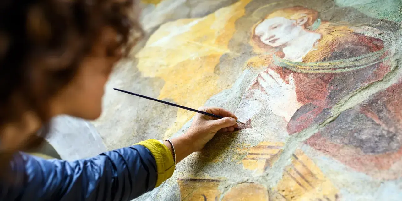 Une artiste restaure une fresque ancienne, sa main tenant un pinceau pour la préparation peinture. La scène montre une figure religieuse aux mains jointes.