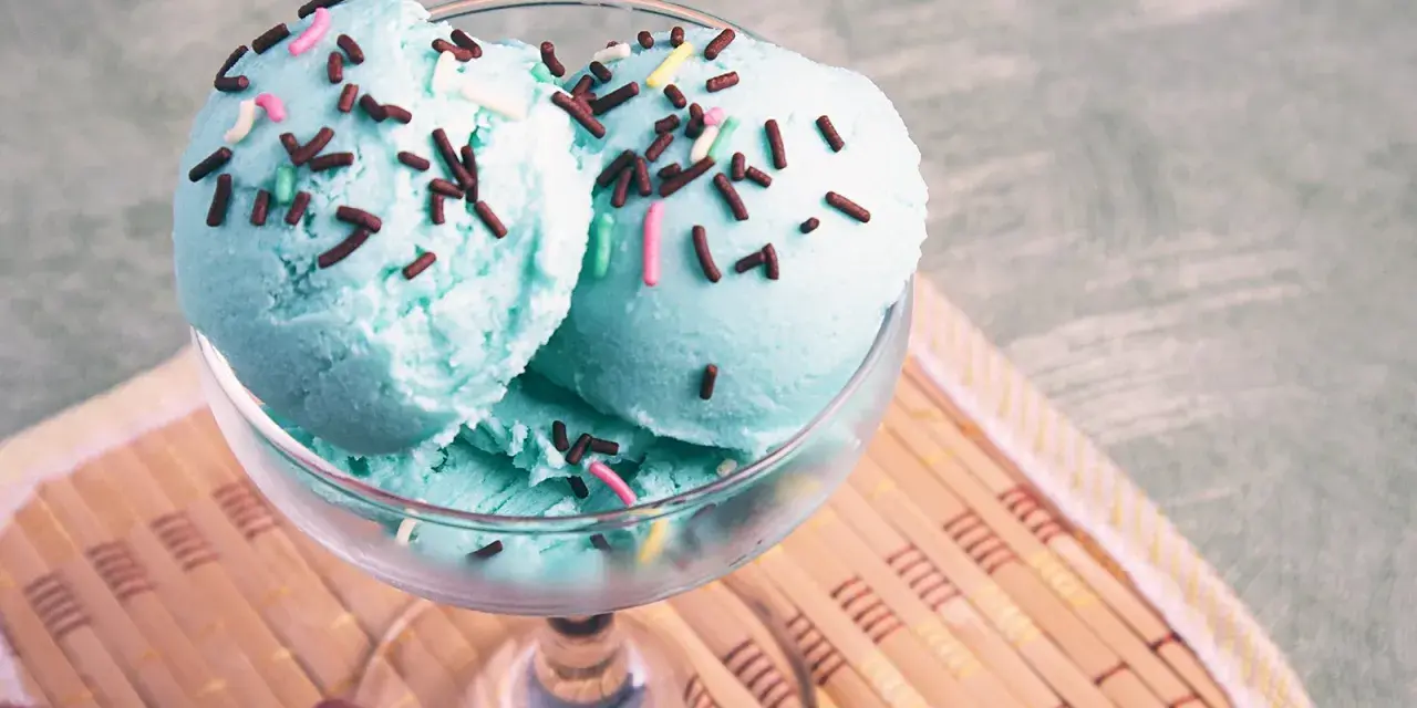 Ein Glas mit blauem Smurf-Eis, bestreut mit bunten Streuseln, steht auf einem Holzuntersetzer.