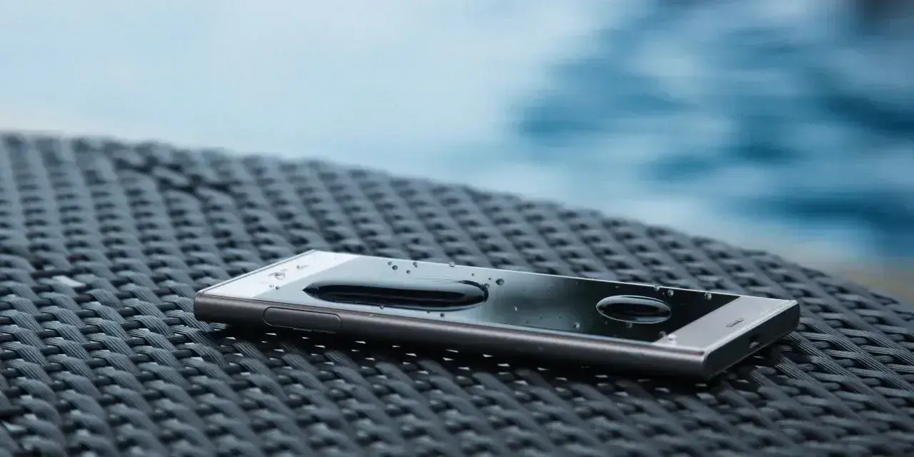 Smartfon z kroplami wody na ekranie, leżący na plecionej powierzchni. W tle widać basen. IP68 co oznacza? Ochronę przed wodą i pyłem.