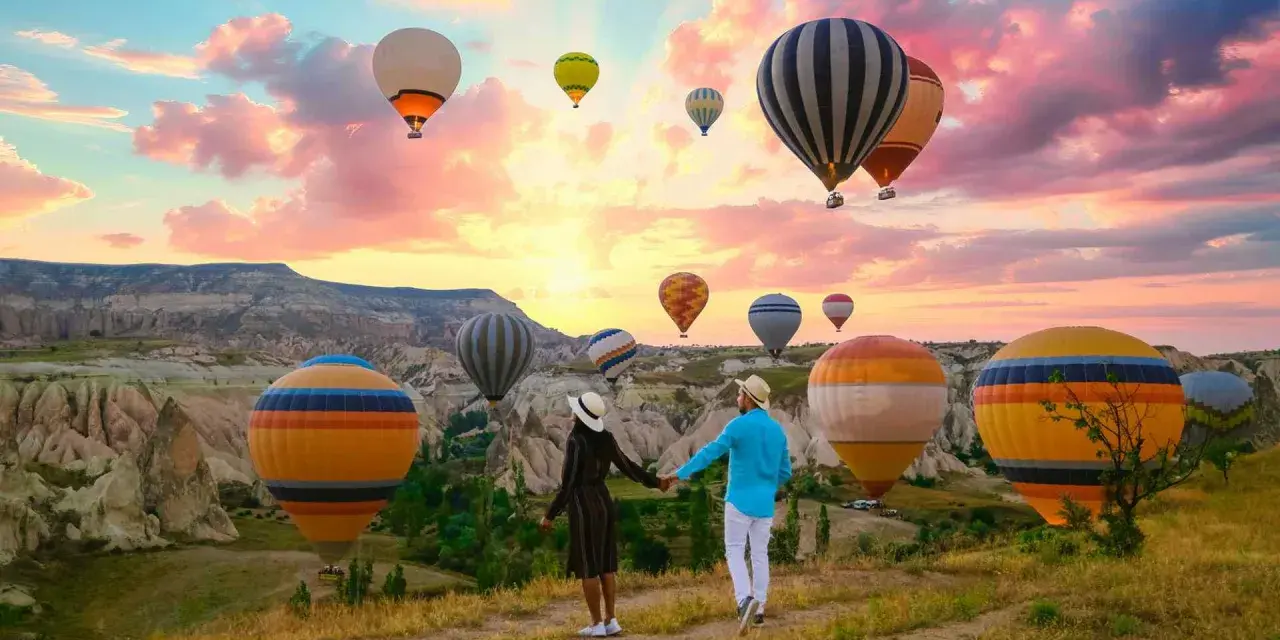 A couple holds hands, gazing at a sky filled with hot air balloons over the unique landscape of western Turkey at sunrise.