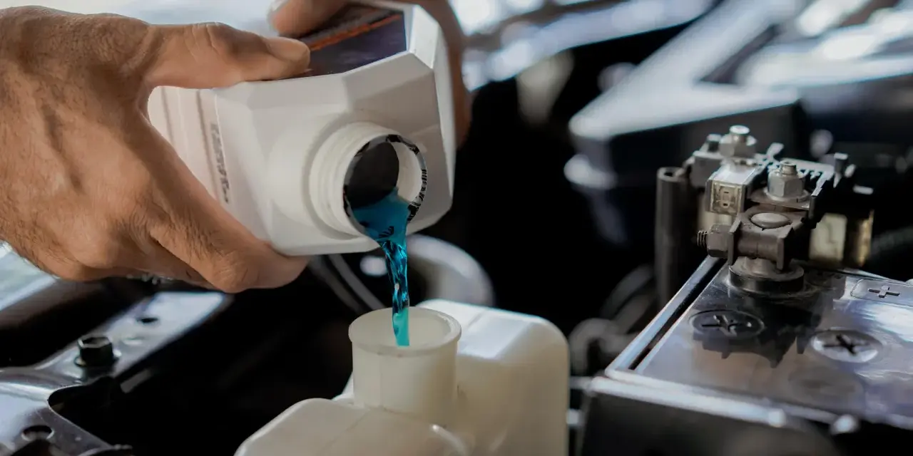 Hands pour blue coolant into a car's reservoir. This maintenance task is essential for preventing overheating, which can lead to costly car radiator replacement.