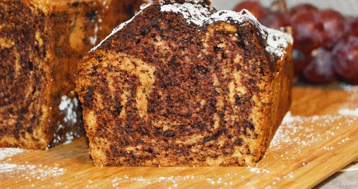 Puszysty chlebek bananowy z kakao, oprószony cukrem pudrem, prezentuje się apetycznie na drewnianej desce.