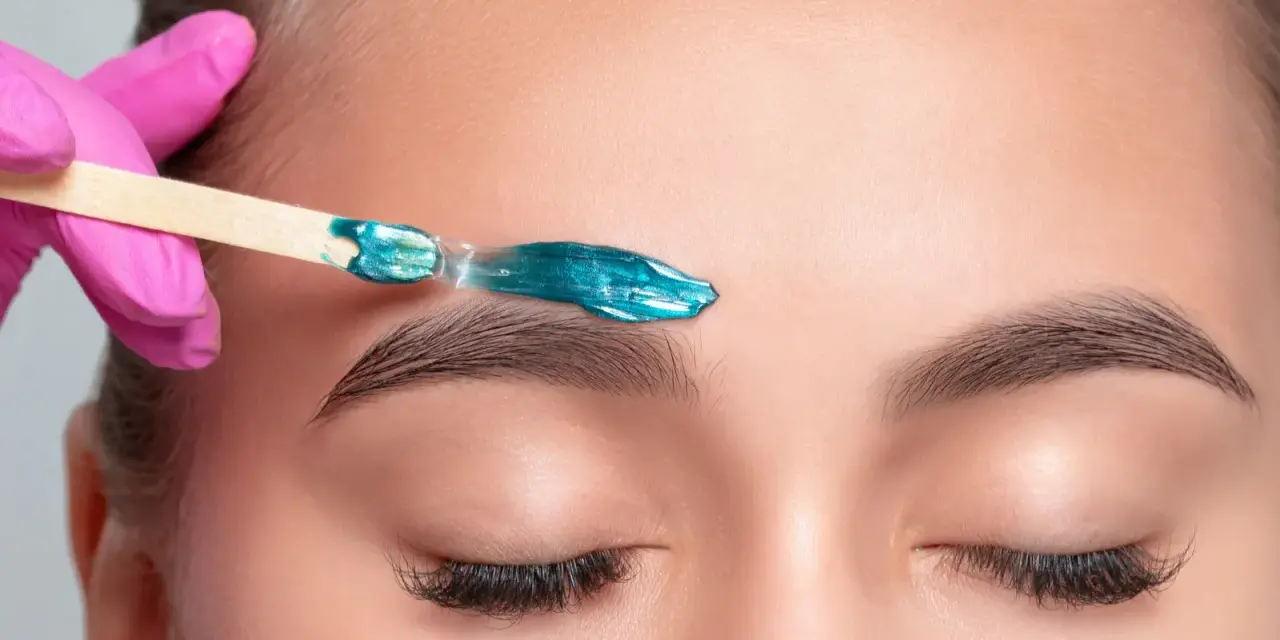 A pink-gloved hand applies turquoise wax to an eyebrow. This close-up shows the contrast between hot wax vs cold wax application for brow shaping.
