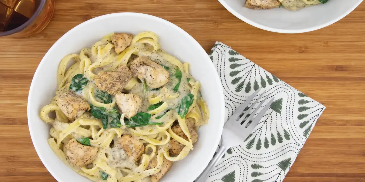 Delicioso plato de pasta alfredo con trozos de pollo y espinacas, servido en un tazón blanco sobre una tabla de madera.