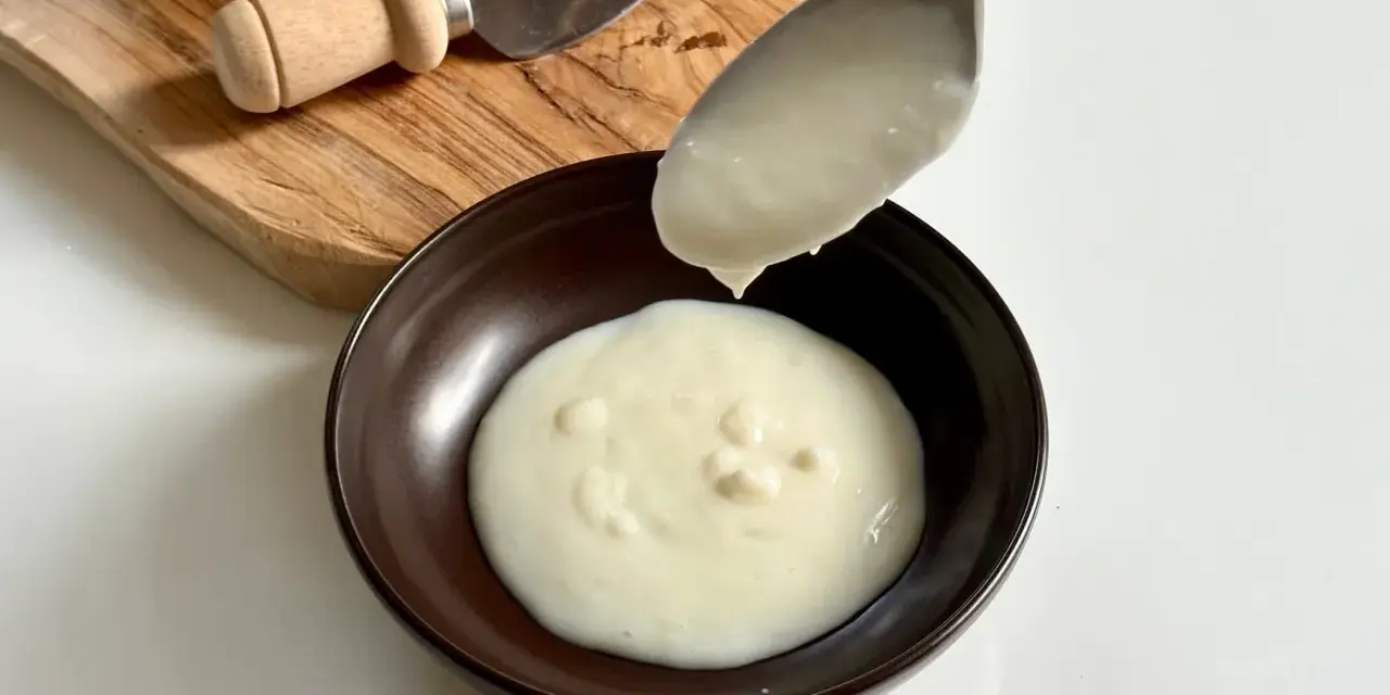 Una cucchiaiata di cremosa fonduta di parmigiano cade in una ciotola. Sul tagliere, un pezzo di Parmigiano Reggiano e un coltellino.
