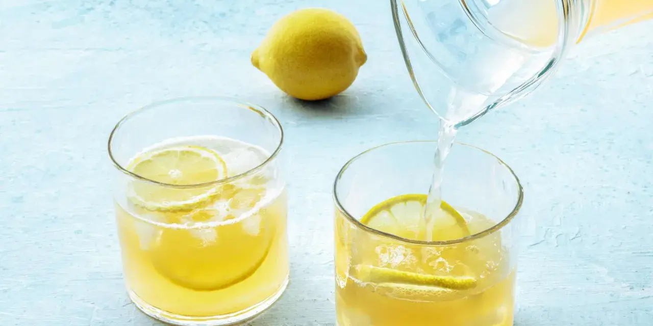 Erfrischendes zitronenwasser am morgen wird in ein Glas mit Eis und Zitronenscheiben gegossen. Eine ganze Zitrone liegt daneben.
