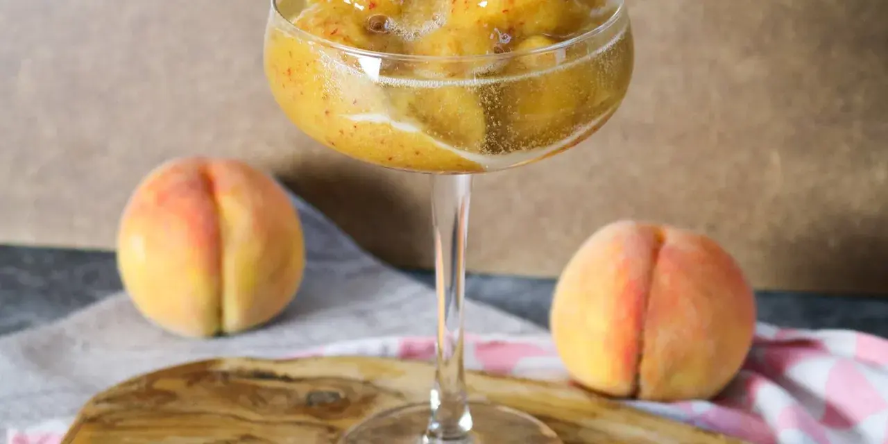 Erfrischendes Pfirsich-Prosecco-Sorbet in einem eleganten Glas, daneben reife Pfirsiche.