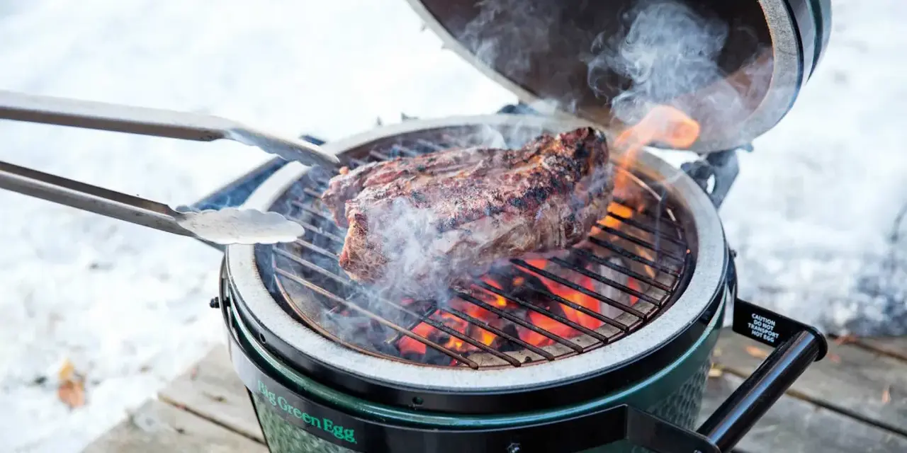 Un succulento bistec a la fiorentina cuoce sulla griglia di un Big Green Egg, circondato da fumo e fiamme.