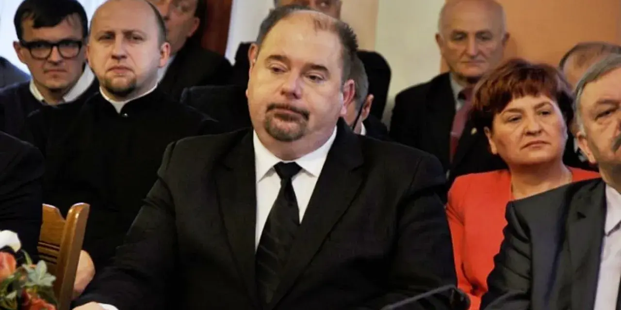 Jarosław Paluch w czarnym garniturze siedzi przy stole, otoczony innymi osobami.