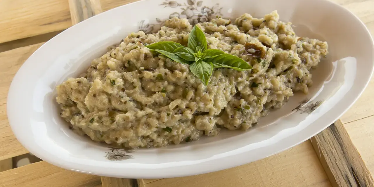 Hummus di melanzane cremoso, guarnito con basilico fresco, servito in un piatto ovale su un tavolo di legno grezzo.