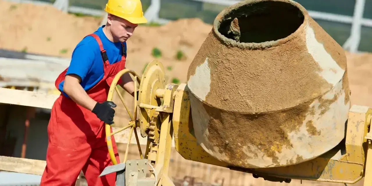 Robotnik w żółtym kasku i czerwonym ogrodniczkach obsługuje betoniarkę. Mieszanka cementowa wygląda na gładką, bez widocznego żwiru, co sugeruje specyficzne proporcje betonu.