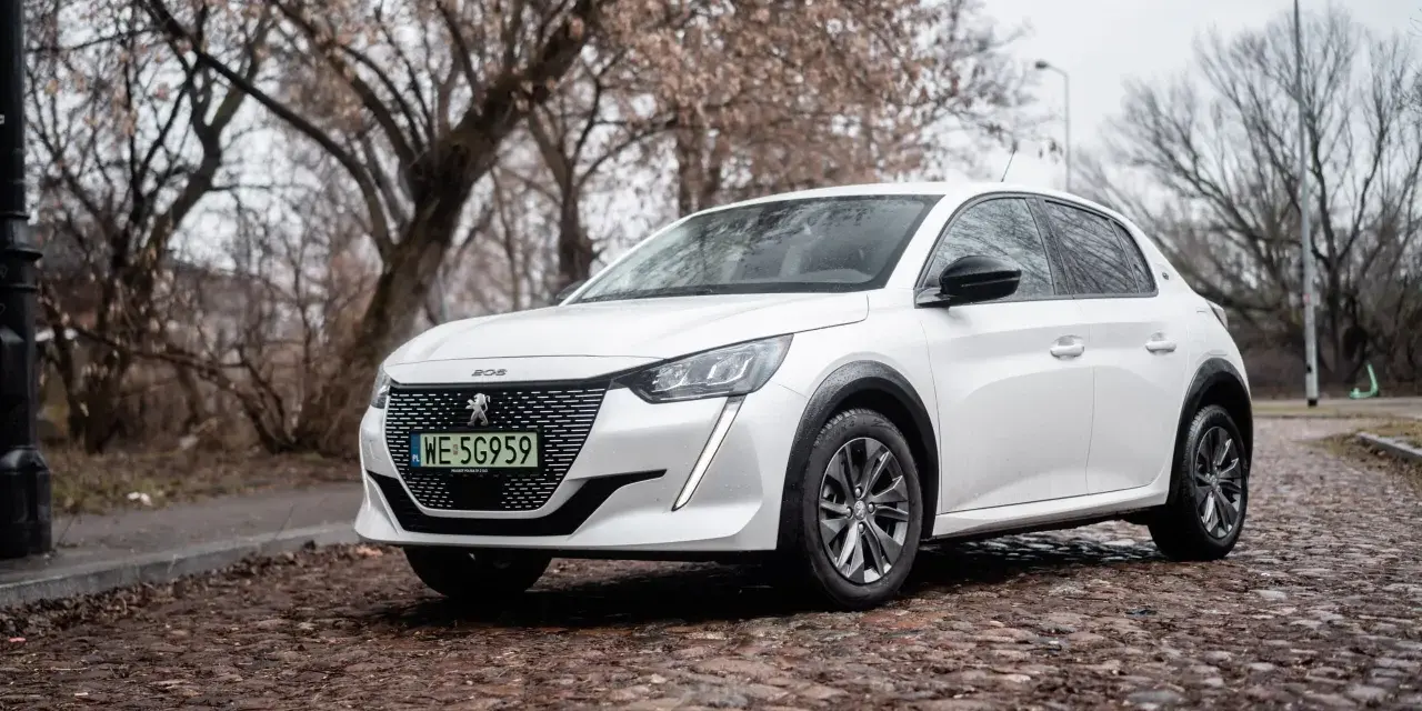 Biały Peugeot e-208 na brukowanej ulicy, z drzewami w tle.