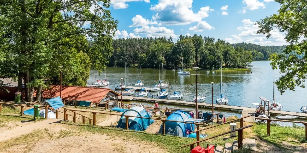 Mazury pod namiot: namioty i żaglówki przy pomoście, zielone drzewa i błękitne niebo z chmurami.