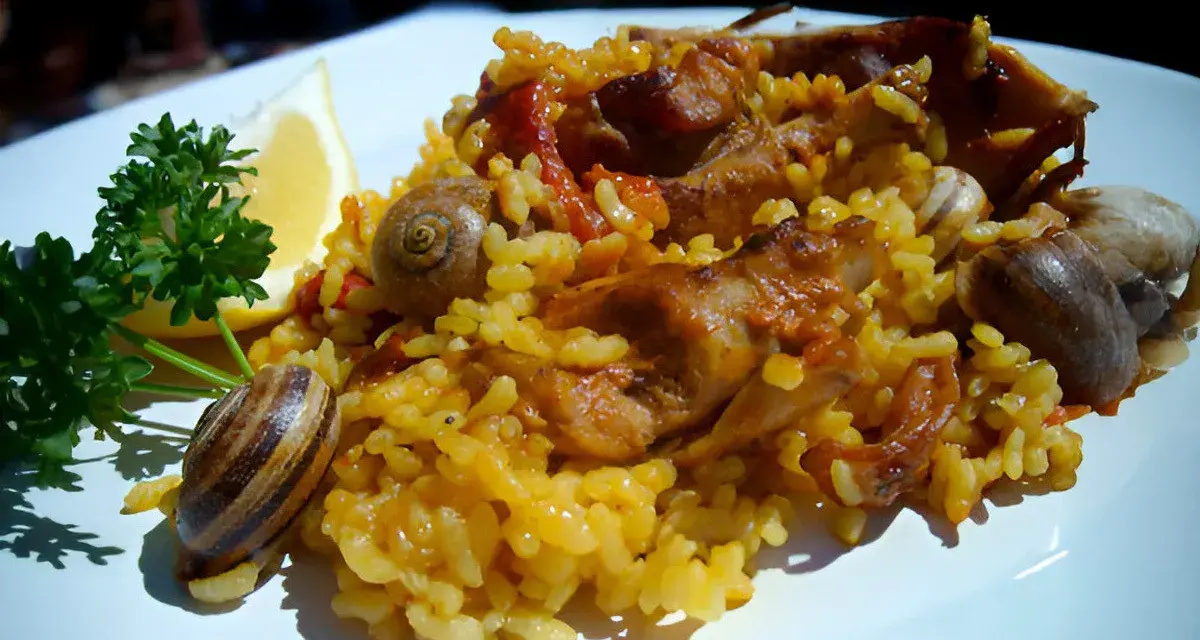 Arroz con conejo, un plato tradicional con trozos de carne dorada, arroz amarillo y caracoles, adornado con perejil y limón.