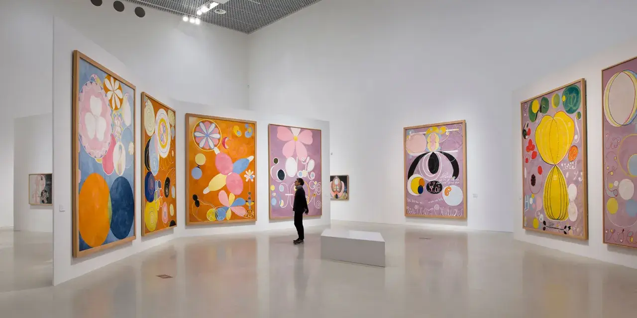 En person betraktar en samling färgstarka, abstrakta målningar av Hilma af Klint i ett ljust galleri.