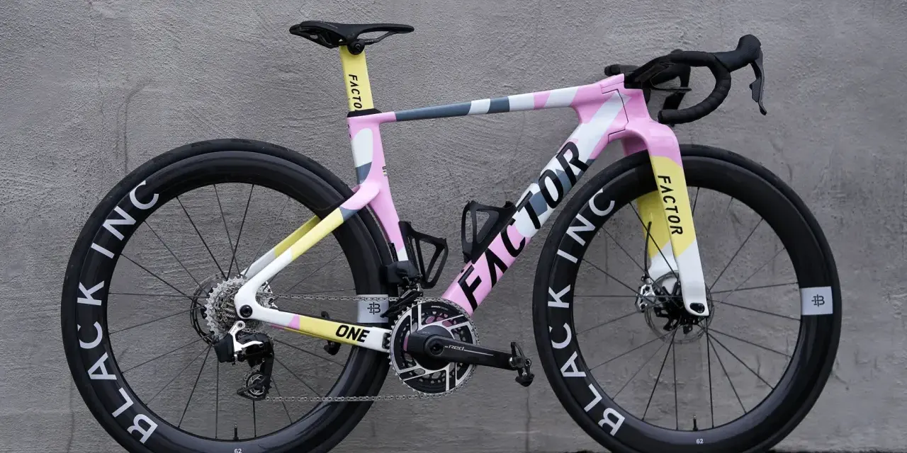 A pink, yellow, and gray Factor road bike with black wheels against a gray wall.