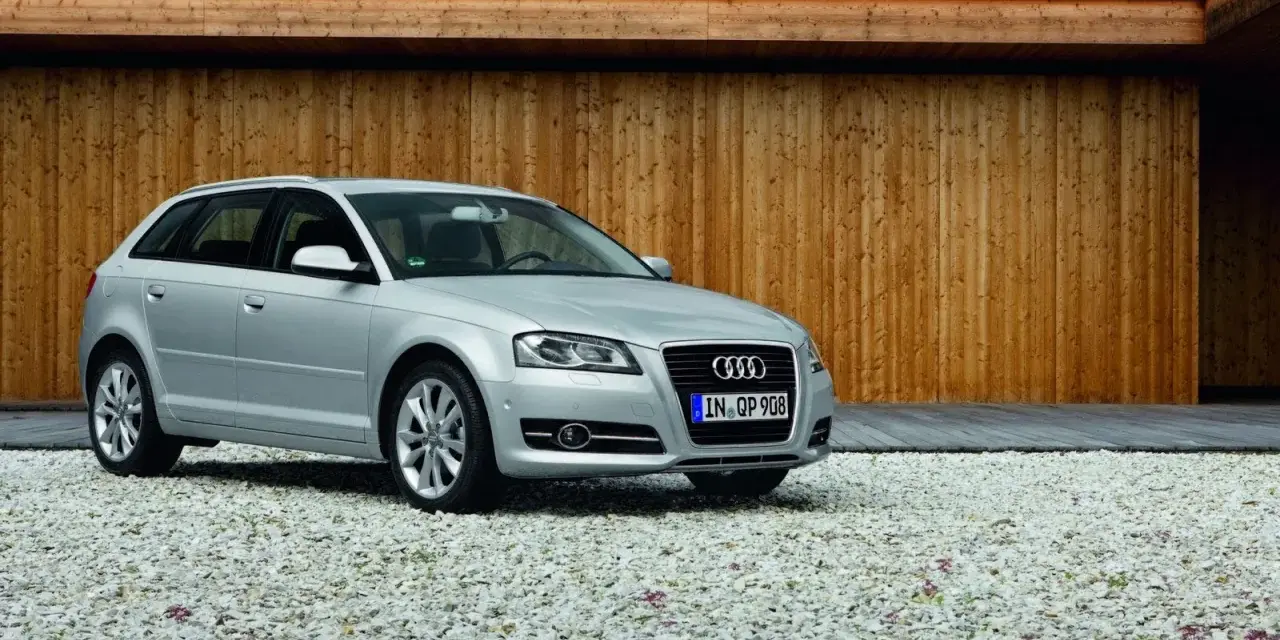 Silberner Audi A3 Baujahr 2008 steht auf einem Kiesparkplatz vor einer Holzwand.