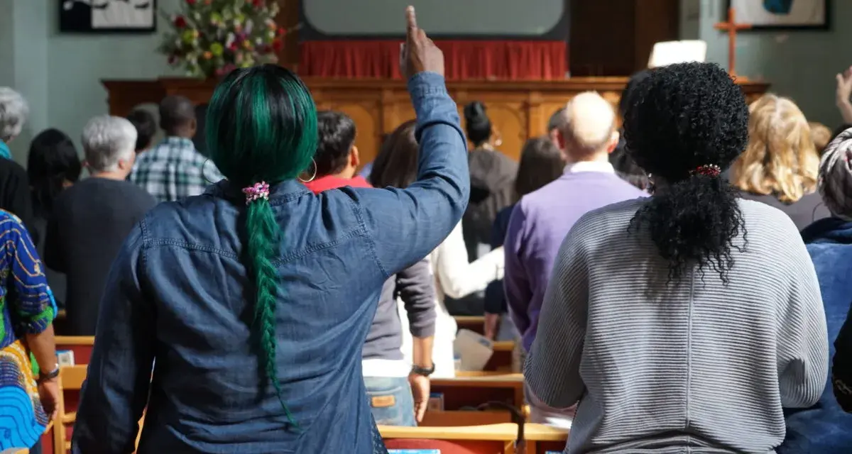 A woman with green hair raises her hand, perhaps inspired by the lyrics on screen, offering chant ideas for the congregation.