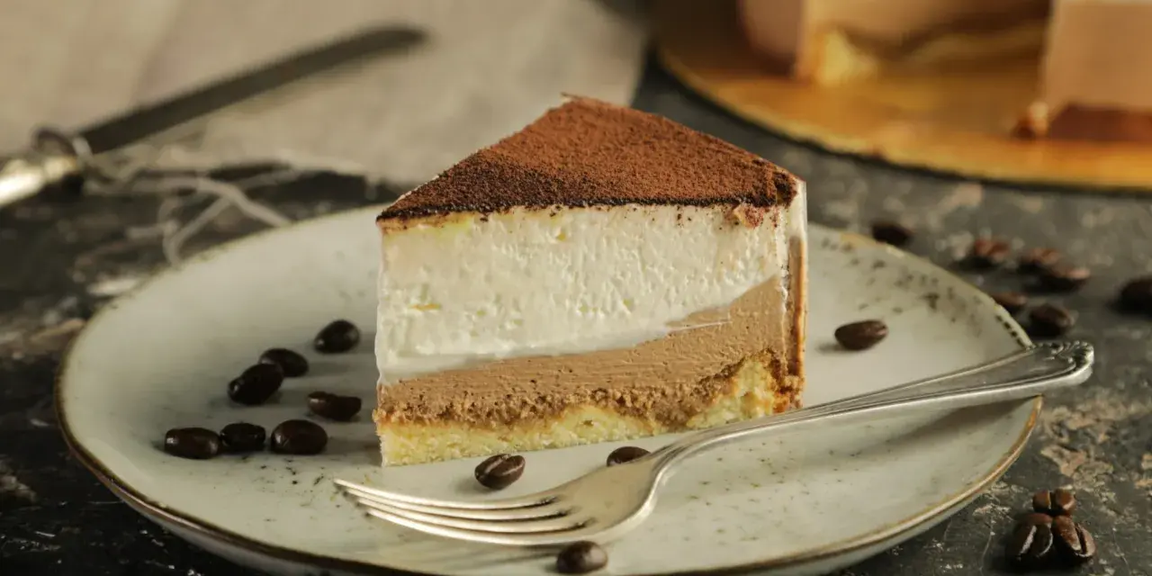 Pyszny sernik tiramisu z warstwami kremu i biszkoptu, posypany kakao. Na talerzu ziarna kawy i widelec.