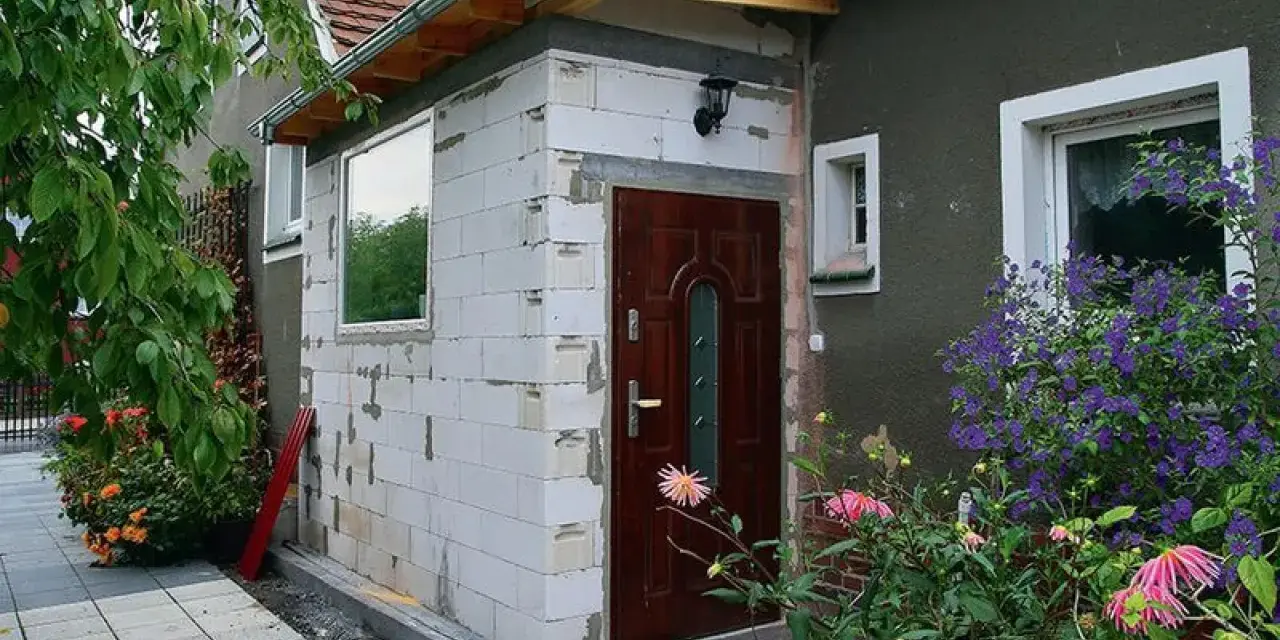 Trwa rozbudowa budynku gospodarczego. Nowa część z białych pustaków, brązowe drzwi, okno i zielone rośliny.