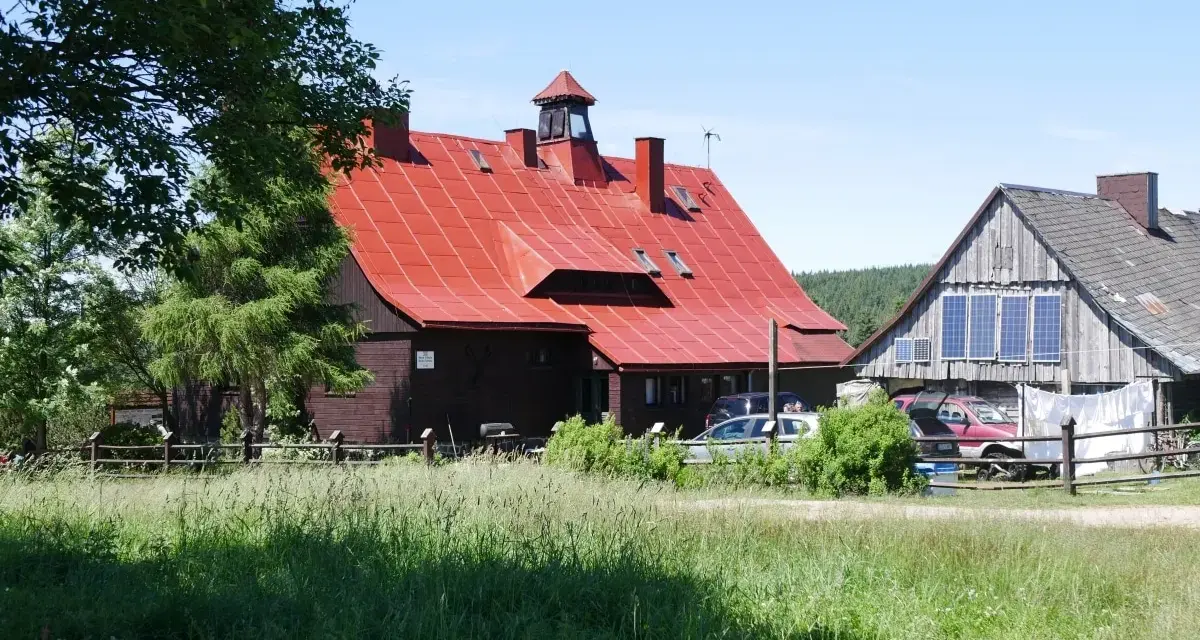 Górzyste krajobrazy, gdzie króluje chatka górzystów z czerwoną dachem. Wokół zieleń i kilka samochodów.