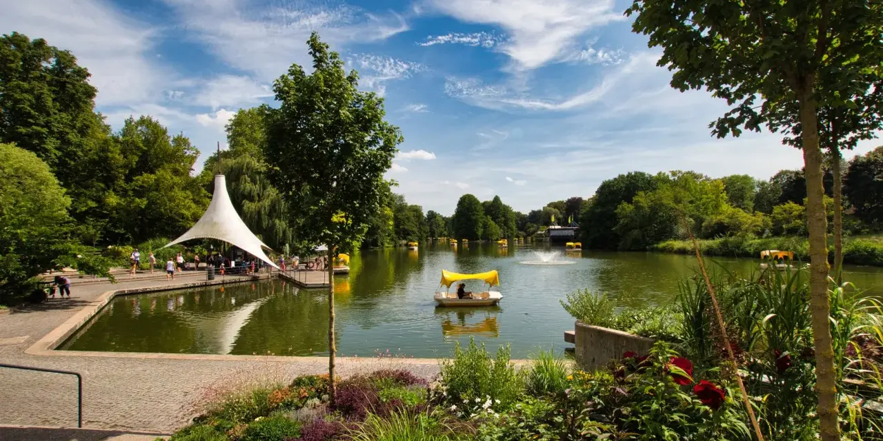 Ein sonniger Tag am Badesee Mannheim. Mehrere Tretboote mit gelben Verdecken gleiten über das Wasser, umgeben von üppigem Grün und blühenden Blumen.