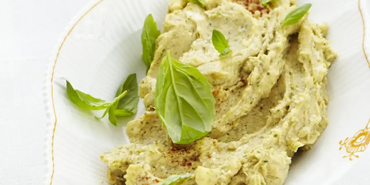 Herzhafte Brotaufstriche selber machen: Cremiger Hummus mit frischem Basilikum und Paprikapulver garniert, serviert in einer ovalen Schale.