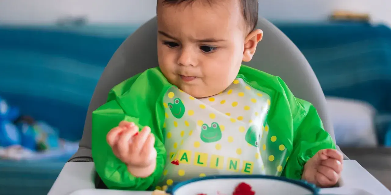 Bebé con babero verde, aprendiendo a comer con las manos. En su plato hay trozos de fruta.